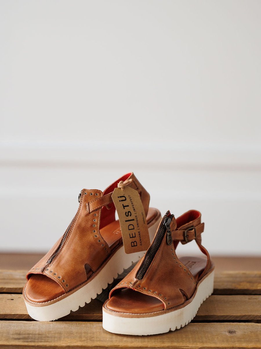 A pair of The Tayten brown distressed leather platform sandals by Savannah Sevens western life{&}style, featuring statement front zippers and a tag, rest on a wooden surface against a plain, softly lit background.