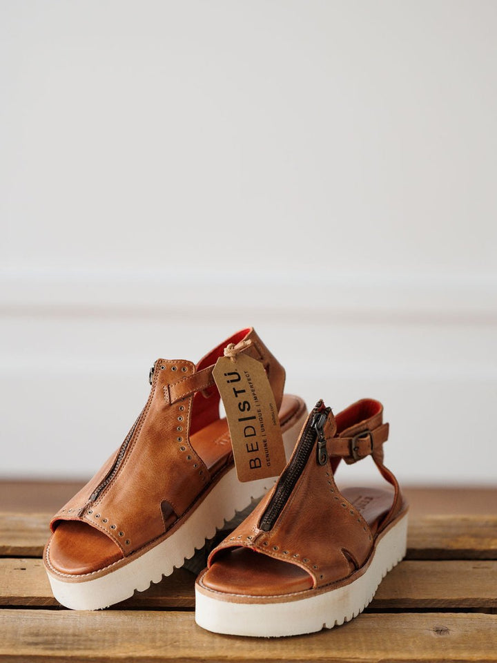 A pair of The Tayten brown distressed leather platform sandals by Savannah Sevens western life{&}style, featuring statement front zippers and a tag, rest on a wooden surface against a plain, softly lit background.