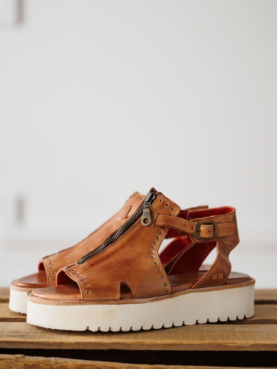 The Tayten by Savannah Sevens western life{&}style: brown statement zipper sandals with studs, white platform soles, and a buckled ankle strap shown on a wooden surface against a light background.