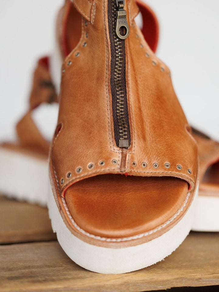A close-up of The Tayten by Savannah Sevens western life{&}style: a statement zipper sandal in distressed brown leather with metal rivets and a thick white platform sole, displayed on a wooden surface. Sandals made by BedStu.