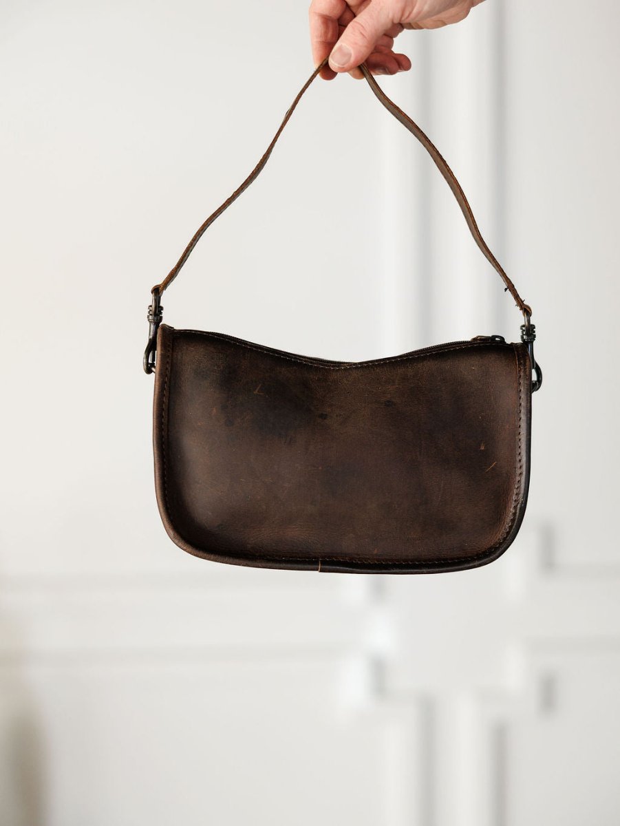 A hand holds up The Texas City, a small dark brown leather crossbody bag by Savannah Sevens western life{&}style, with a single strap against a light, blurred background.
