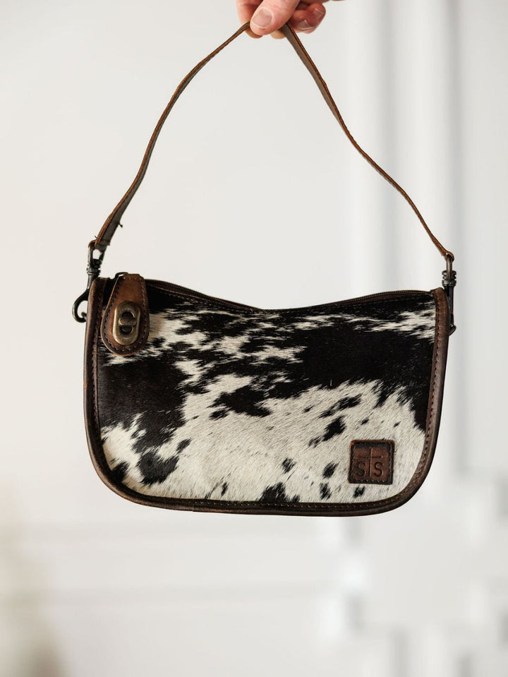 A hand holds The Texas City leather crossbody bag by Savannah Sevens western life{&}style, featuring a brown strap and trim, black and white cowhide pattern, and a small logo patch on the front.