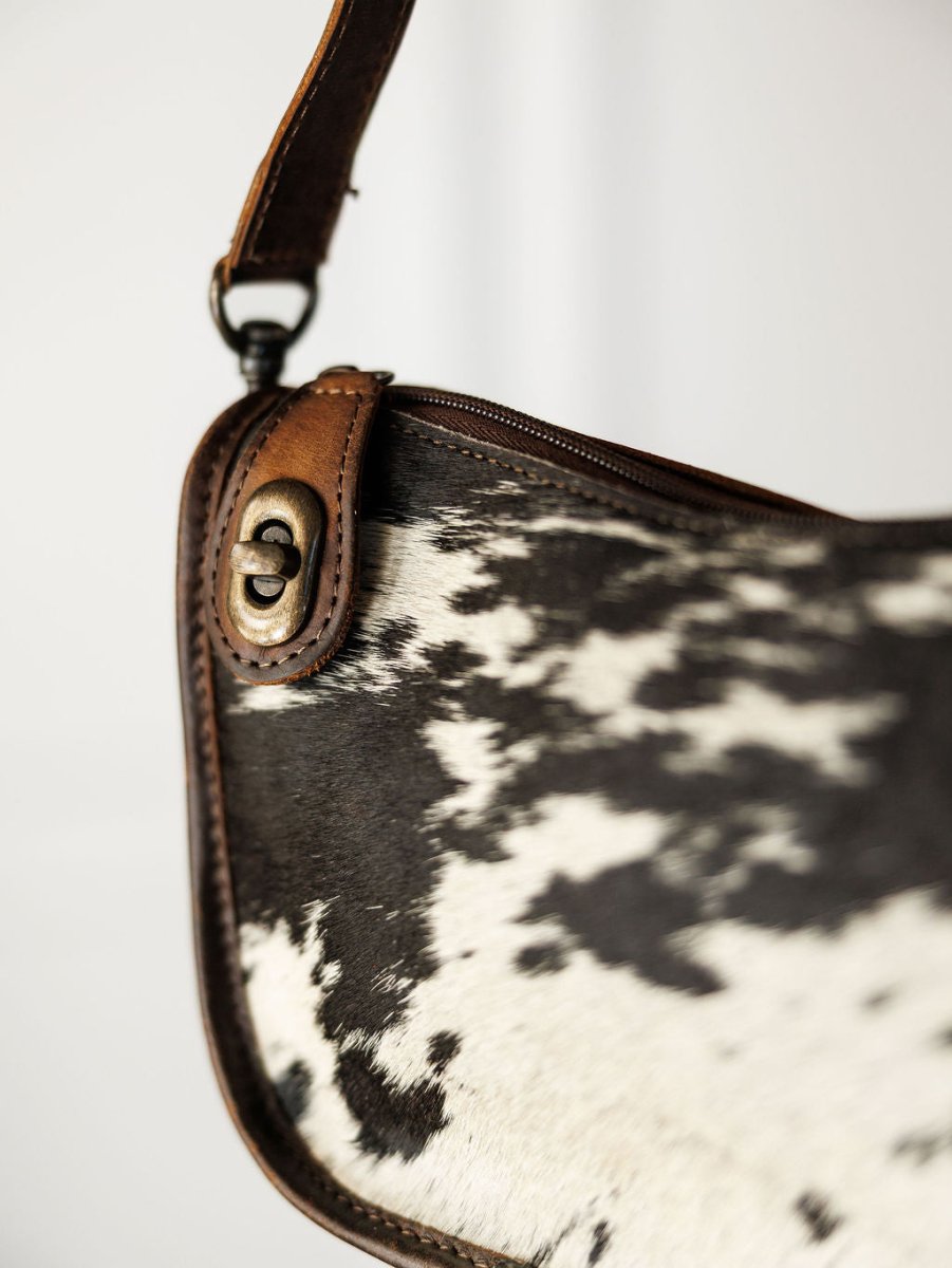 Close-up of The Texas City cowhide crossbody bag by Savannah Sevens western life{&}style, featuring a brown leather strap and trim, black and white pattern, and metal clasp detail.