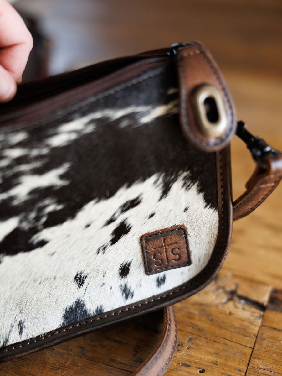 A close-up of The Texas City cowhide crossbody bag by Savannah Sevens western life{&}style, featuring brown leather edges and a stitched leather patch with "s|s", resting on a wooden surface.