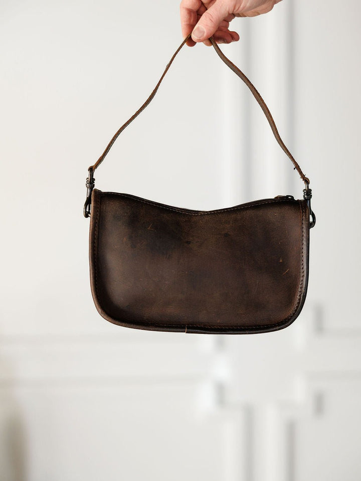 A hand holds up The Texas City, a small dark brown leather crossbody bag by Savannah Sevens western life{&}style, with a single strap against a light, blurred background.