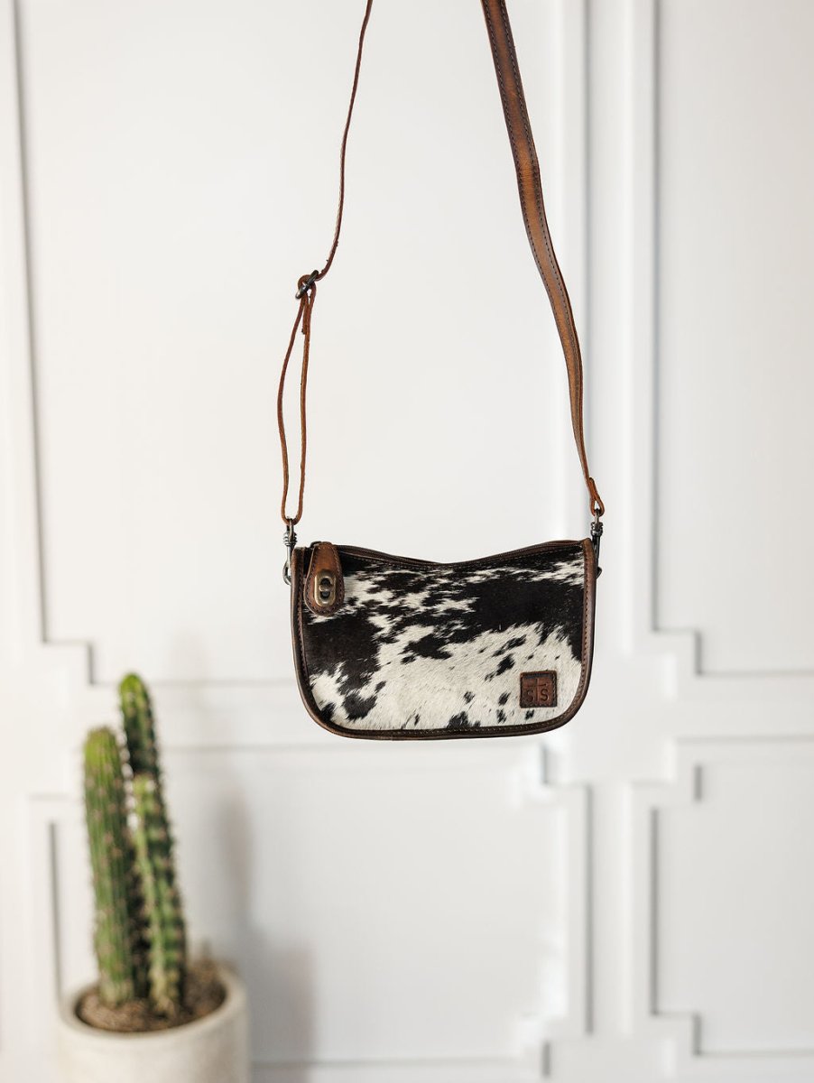 The Texas City cowhide crossbody bag from Savannah Sevens western life{&}style hangs against a white wall, with a potted cactus visible in the lower left corner.