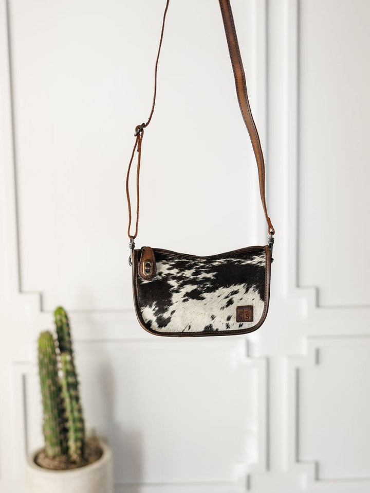 The Texas City cowhide crossbody bag from Savannah Sevens western life{&}style hangs against a white wall, with a potted cactus visible in the lower left corner.
