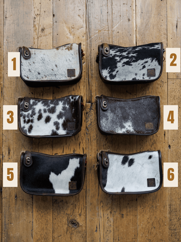 Six The Texas City wallets by Savannah Sevens western life{&}style, each with a unique black and white cowhide pattern, are arranged in two rows on a wooden surface and labeled with numbers 1 to 6.