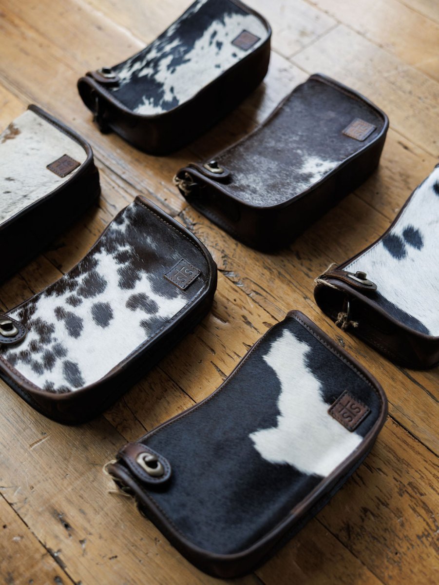 Six The Texas City cowhide crossbody bags by Savannah Sevens western life{&}style, in black, white, and brown patterns, are arranged on a wooden floor. Each bag features unique markings and a metal ring on one corner.