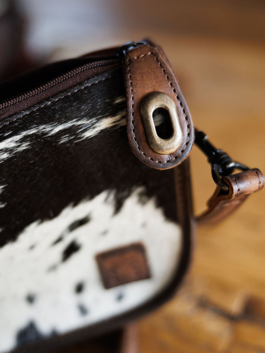Close-up of The Texas City cowhide crossbody bag by Savannah Sevens western life{&}style, featuring black and white fur, a brown leather strap, brass hardware, and visible stitching details on a wooden surface.