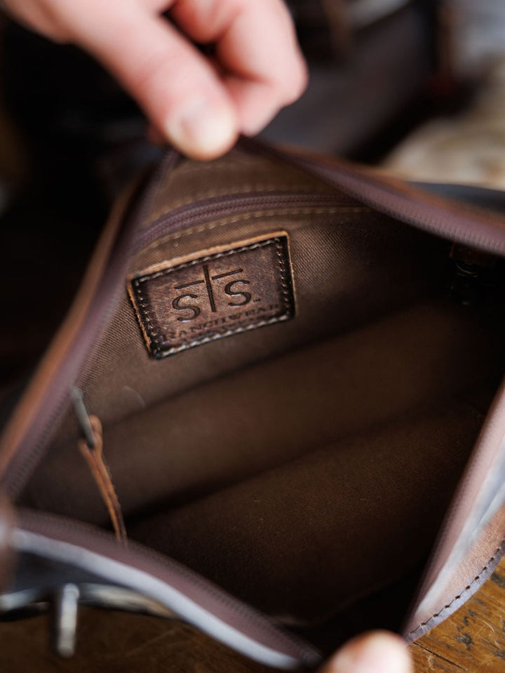A close-up of hands holding open The Texas City brown leather crossbody bag by Savannah Sevens western life{&}style, showing the inside lining and a stitched leather label with s|s embossed on it.