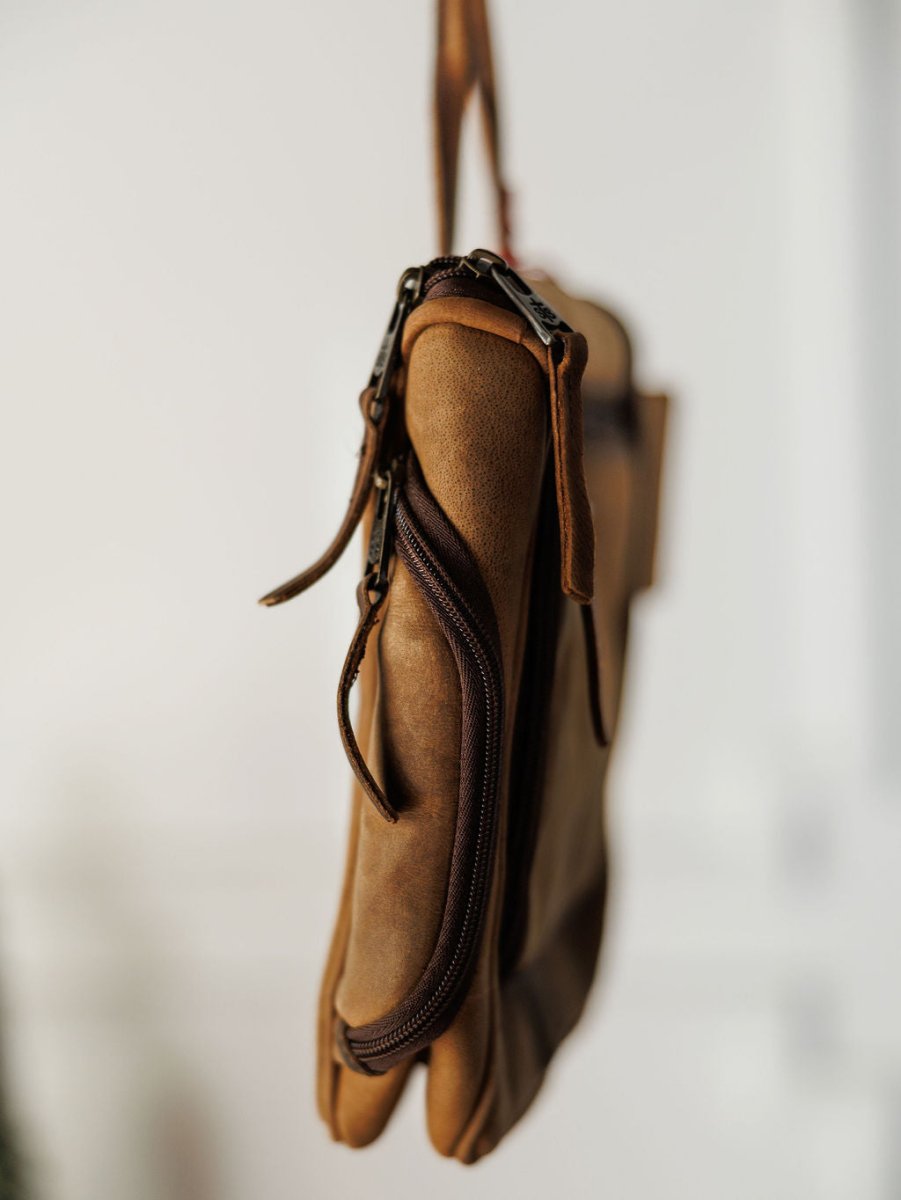 A close-up side view of Savannah Sevens western life{&}style's The Toledo, a brown leather hanging toiletry bag with a visible zipper pocket and sturdy strap, set against a blurred light background.
