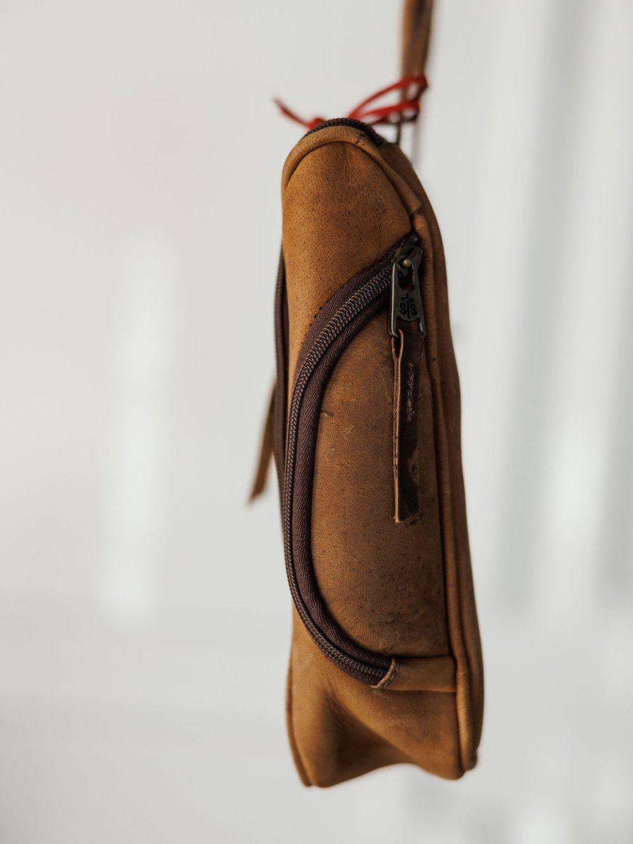 The Toledo shave kit by Savannah Sevens western life{&}style, a small brown leather bag with a dark zipper and leather pull tab, hangs by its strap against a plain white background.