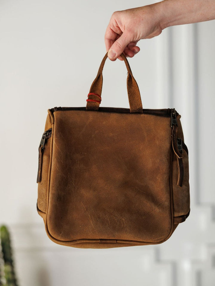 A hand holds The Toledo leather bag by Savannah Sevens western life{&}style against a neutral indoor background. The brown bag, resembling a shave kit, has visible zippers, slight wear, and a red band on one handle.