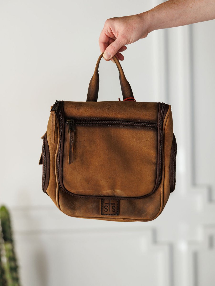 A hand holds up The Toledo, a brown leather shave kit with a top handle and front zipper by Savannah Sevens western life{&}style, against a white background with decorative molding.