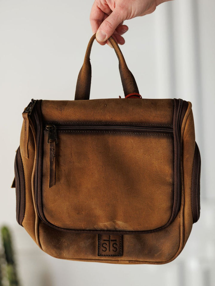 A hand holds up The Toledo leather shave kit by Savannah Sevens western life{&}style, featuring a top handle, front zip pocket, side zippers, and an embossed logo. The softly blurred background highlights this hanging toiletry bag.
