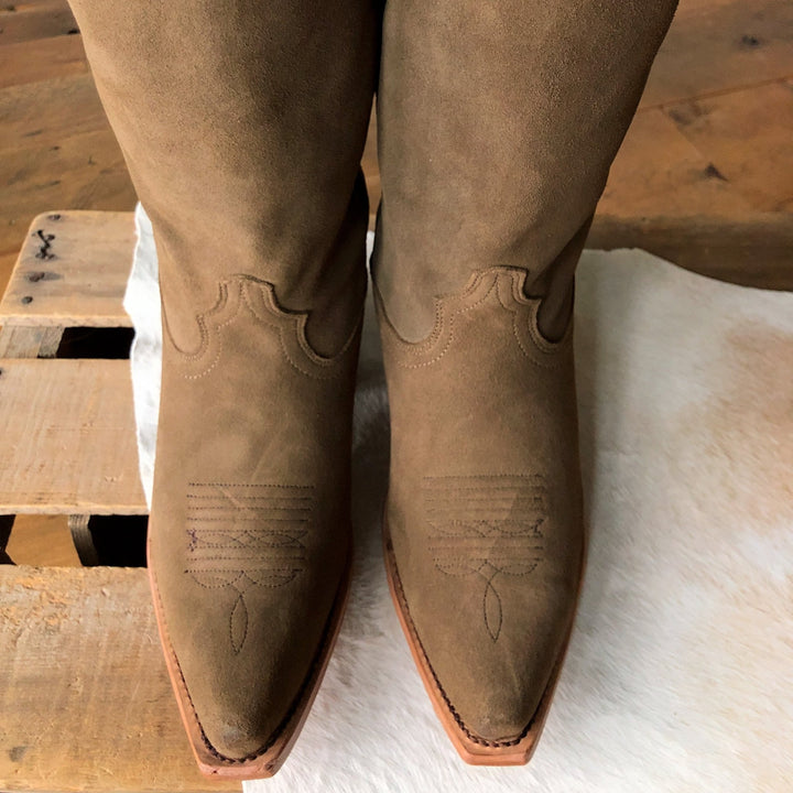 A close-up of The Traver boots by Savannah Sevens western life{&}style, featuring brown rough out leather, snip toes with decorative stitching, displayed on a light hide beside a wooden pallet.Made by Black Star. 