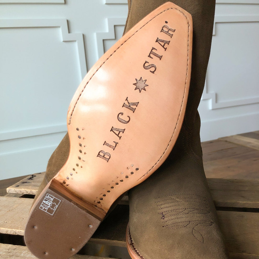 A close-up of The Traver brown snip toe boot by Savannah Sevens western life{&}style shows its light leather sole with the brand name stamped in large letters, resting on a wooden surface before a white paneled wall.