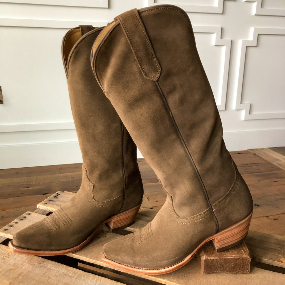 The Traver tan rough out leather boots by Savannah Sevens western life{&}style, featuring wooden heels, are displayed on a wooden surface with a decorative white wall in the background. Made by Black Star. 