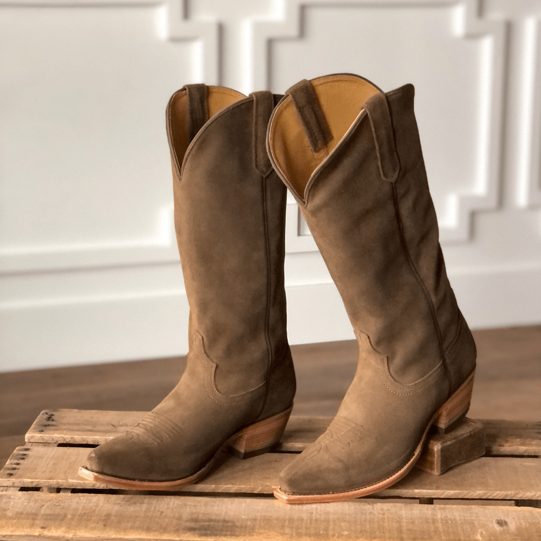 The Traver boots by Savannah Sevens western life{&}style feature brown rough-out leather, snip toes, and angled heels. They are displayed on a wooden pallet with a white decorative wall in the background. Made by Black Star. 