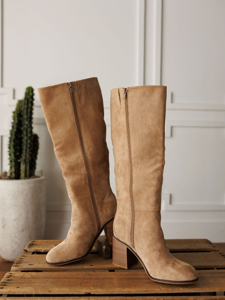 The Tullos knee-high boots by Savannah Sevens western life{&}style, in tan faux suede with round toes, side zippers, and block heels, are displayed on wood with a potted cactus and white panel walls in the background.