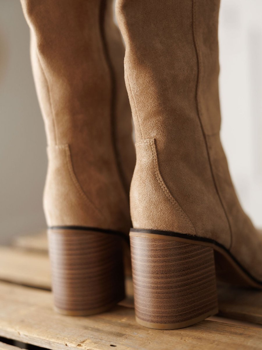 Close-up of The Tullos tan faux suede boots by Savannah Sevens western life{&}style, featuring stacked wooden block heels on a wooden surface. Suede texture and wood grain details are clearly visible.