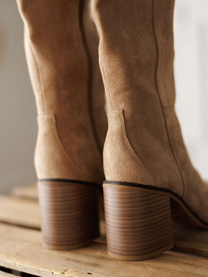 Close-up of The Tullos tan faux suede boots by Savannah Sevens western life{&}style, featuring stacked wooden block heels on a wooden surface. Suede texture and wood grain details are clearly visible.