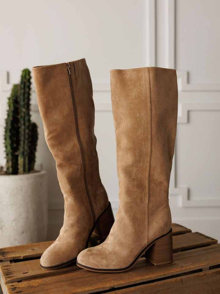 The Tullos by Savannah Sevens western life{&}style: tan faux suede round toe boots with block heels and side zippers, shown on a wooden surface with a potted cactus in the background.