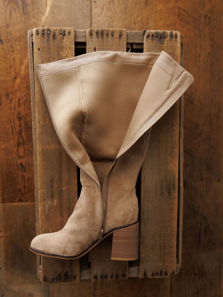The Tullos knee-high boot by Savannah Sevens western life{&}style, in tan faux suede with a chunky heel and side zipper, is shown partially unzipped atop a rustic wooden crate on a brown wooden floor.