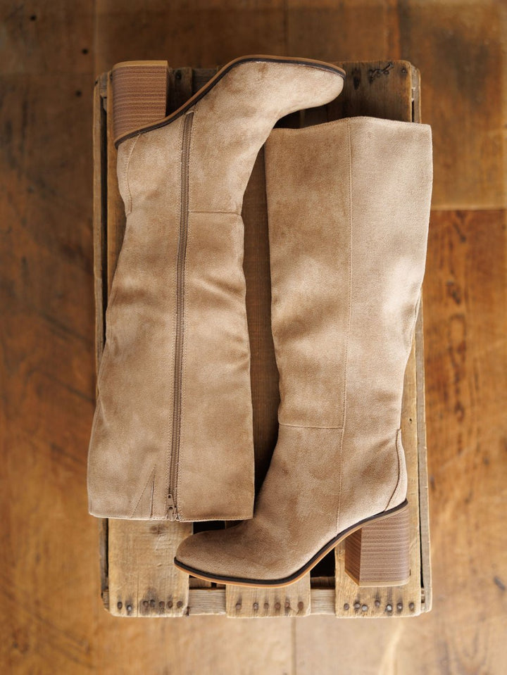 The Tullos by Savannah Sevens western life{&}style: tan faux suede knee-high boots with round toes, block heels, and side zippers, displayed on a wooden crate against a rustic wooden floor.