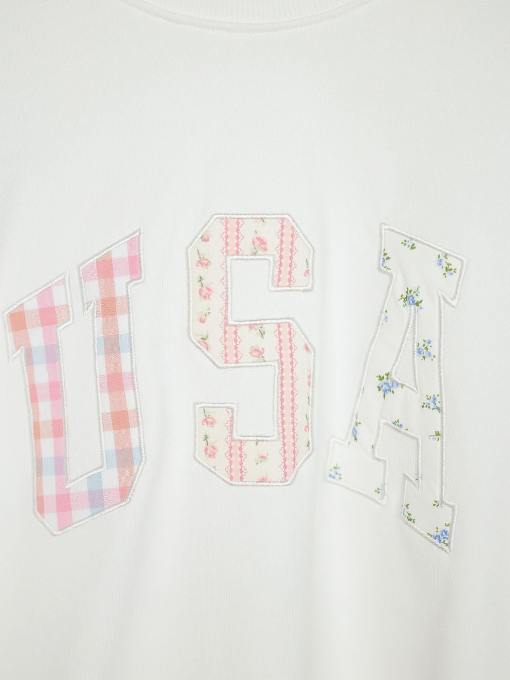 The USA Patchwork Crewneck by Savannah Sevens western life{&}style is a white long sleeve pullover with varsity-style embroidered patchwork letters, each in unique pastel floral or plaid patterns for a charming touch.