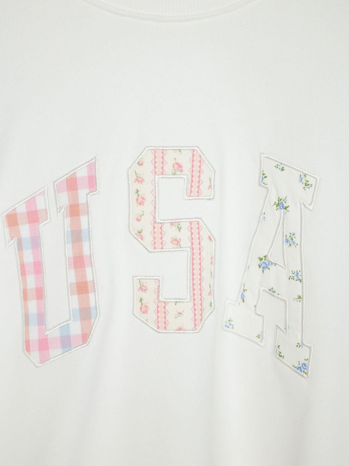 The USA Patchwork Crewneck by Savannah Sevens western life{&}style is a white long sleeve pullover with varsity-style embroidered patchwork letters, each in unique pastel floral or plaid patterns for a charming touch.