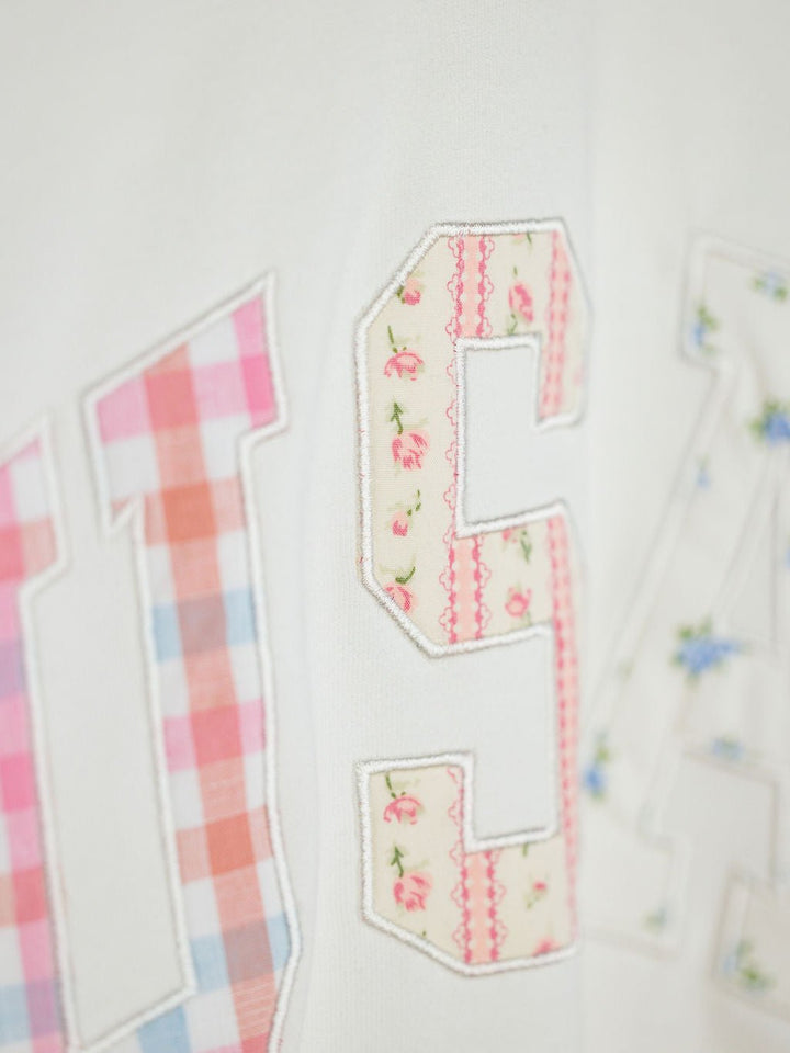 The USA Patchwork Crewneck by Savannah Sevens western life{&}style features embroidered "USA" letters in pink plaid, floral, and blue flower prints stitched on a white long sleeve background.