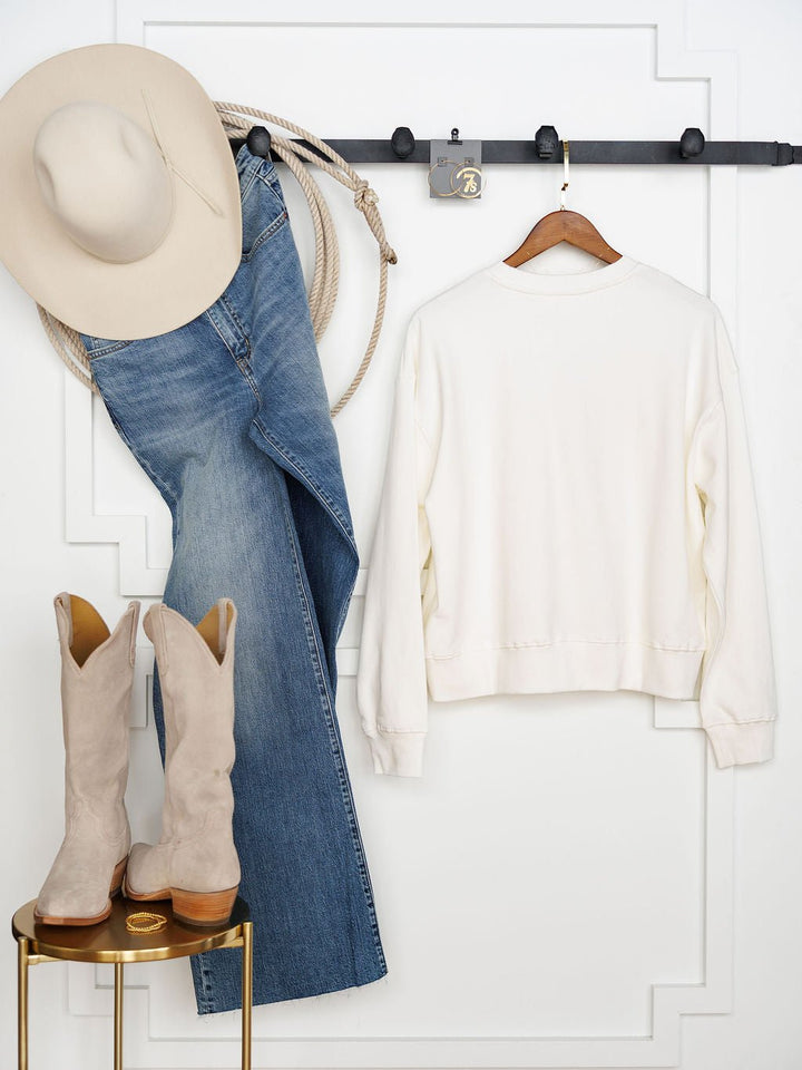 A cream cowboy hat, blue jeans, and a white long sleeve hang on a black rack next to The USA Patchwork Crewneck by Savannah Sevens western life{&}style. Below, beige suede cowboy boots rest on a small round gold table against a white wall.
