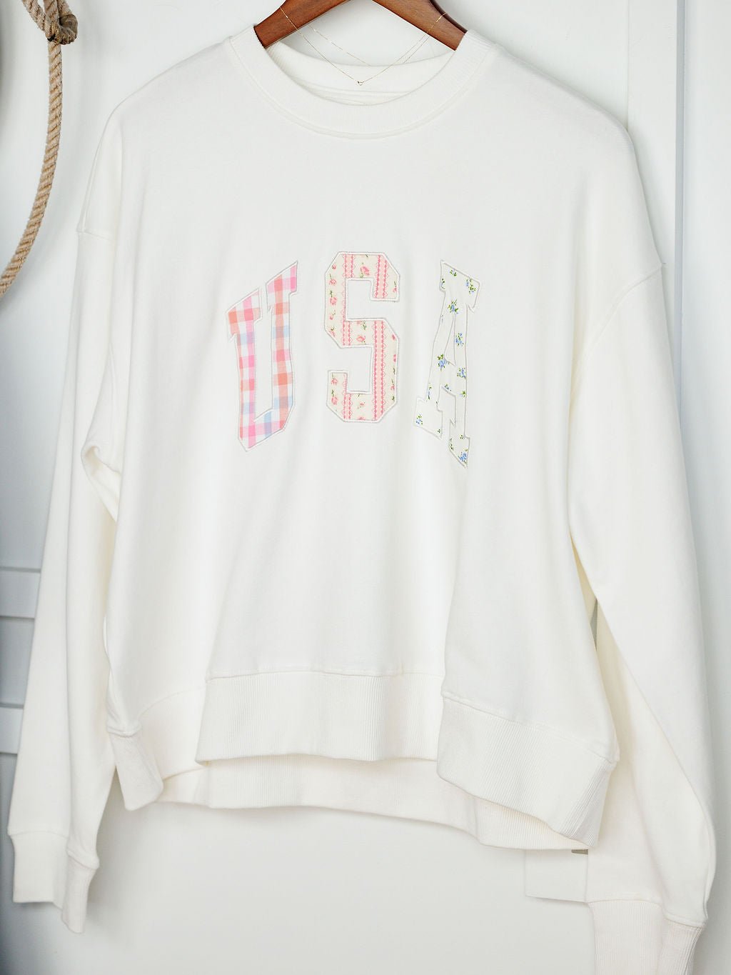 The USA Patchwork Crewneck by Savannah Sevens western life{&}style is a white long sleeve pullover with large pastel embroidered “USA” letters on the front, displayed on a wooden hanger.