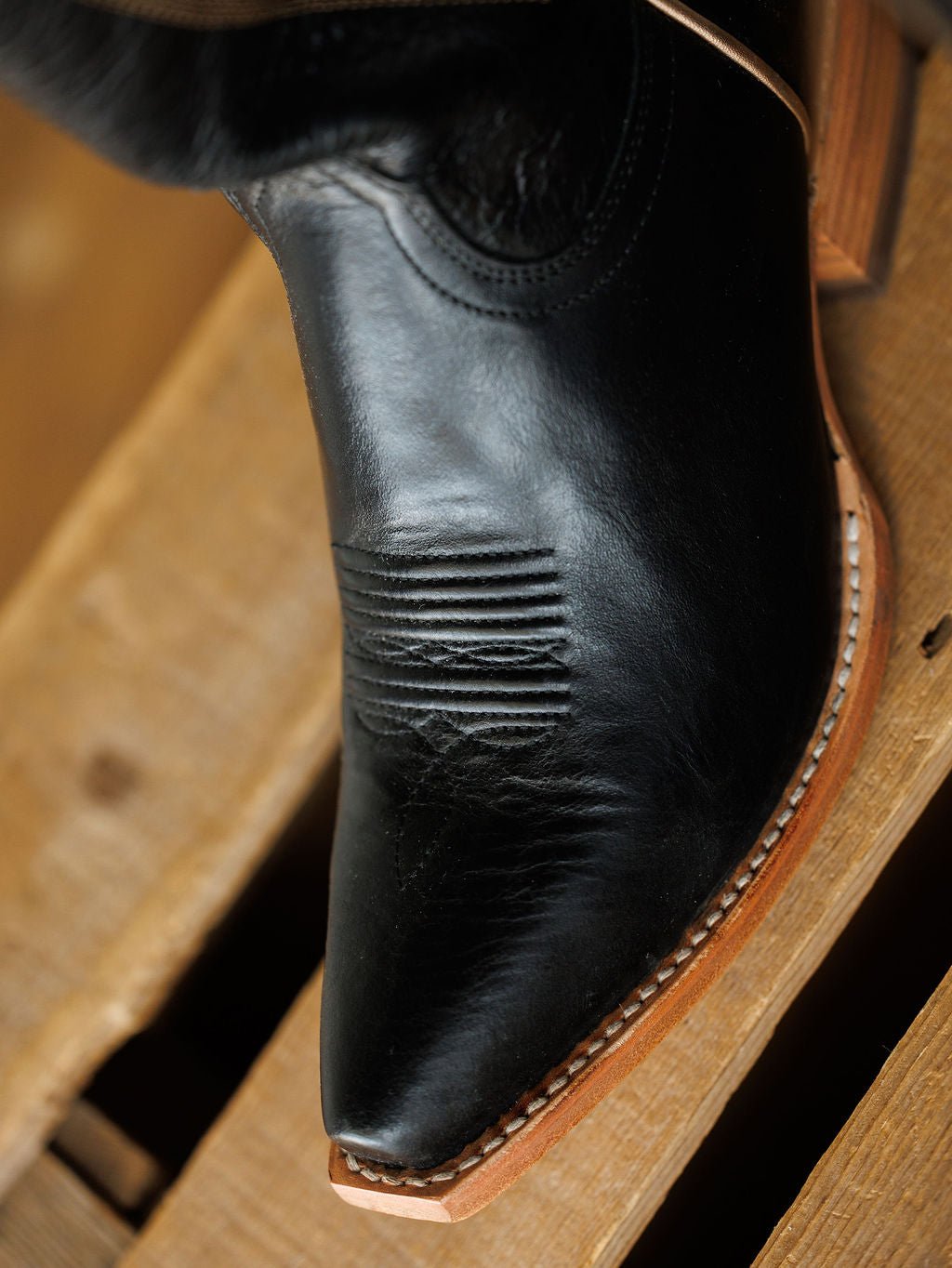 A close-up of The Vaquera boot by Savannah Sevens western life{&}style, featuring black leather, detailed western embroidery, a snip toe, and a light brown sole, resting on wood. Comfortable with a pointed toe design. Made by Black Star. 