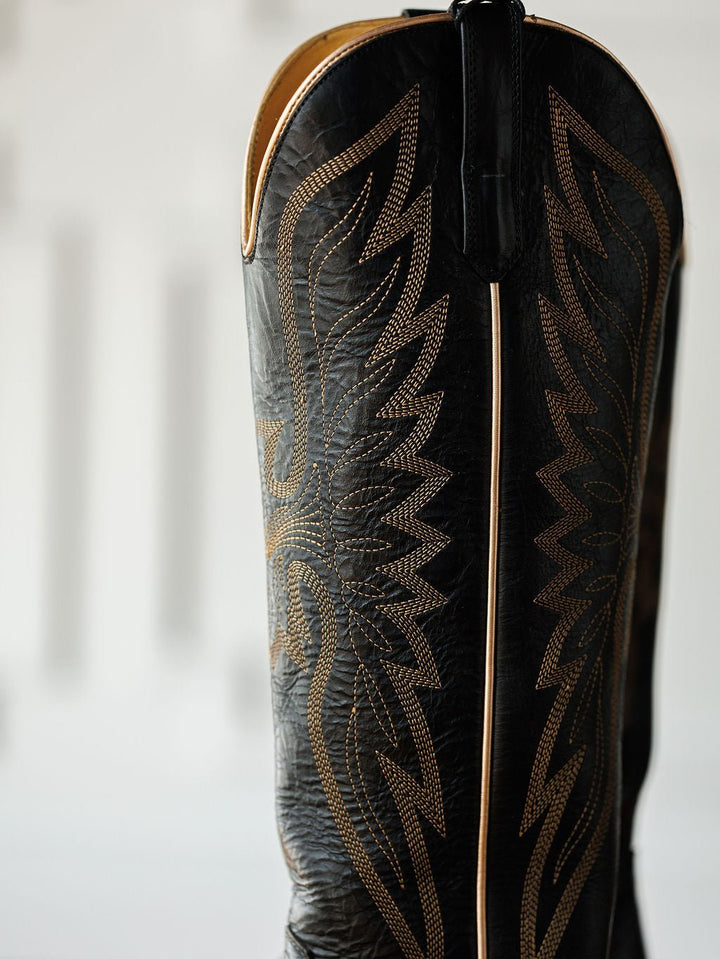 Close-up of The Vaquera by Savannah Sevens western life{&}style—a black leather snip toe boot with golden flame-like western embroidery, vertical seam, and pull strap, shown against a blurred light background. Made by Black Star. 