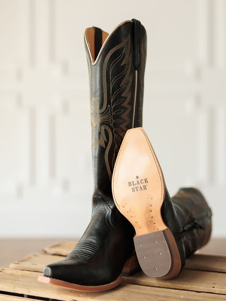 A pair of The Vaquera black leather snip toe boots by Savannah Sevens western life{&}style, with western embroidery; one stands upright, the other on its side showing the tan sole stamped BLACK STAR. Both rest on a wooden surface. Made by Black Star. 