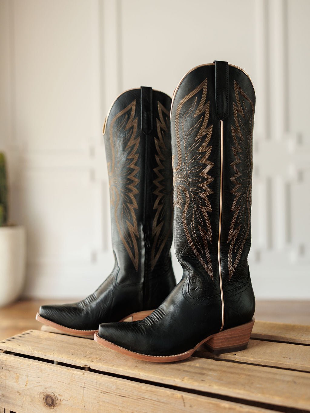The Vaquera by Savannah Sevens western life{&}style: black leather snip toe boots featuring brown stitching and western embroidery, shown on a wooden surface indoors with a blurred background. Made by Black Star. 