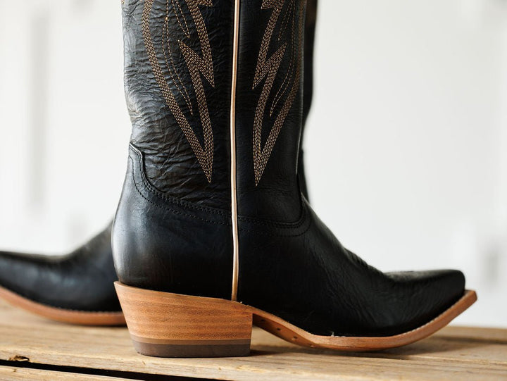 A close-up of The Vaquera by Savannah Sevens western life{&}style—black leather western boots with decorative shaft stitching, a brown stacked heel, and a pointed snip toe—resting on a wooden surface. Made by Black Star. 