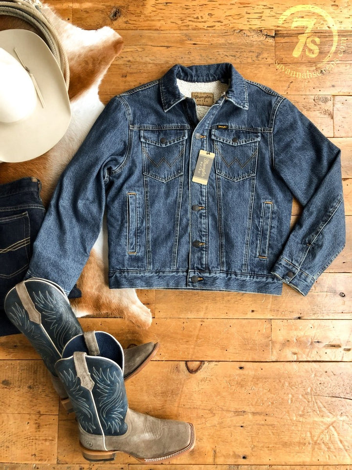 A blue "The Wagoner" men's jacket by Savannah Sevens western life&style with a tag is on a wooden floor beside cowboy boots, dark jeans, a white cowboy hat, and a cowhide rug. The 7s logo appears in the top right corner.