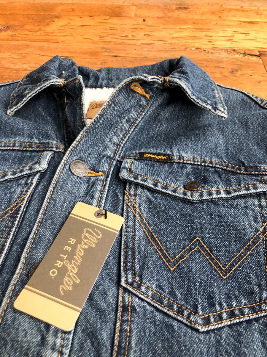 A close-up of The Wagoner Men's jacket by Savannah Sevens western life{&}style—blue denim with tan stitching, metal buttons, and a Wrangler Retro tag—laid on wood for an iconic men's button-up style.