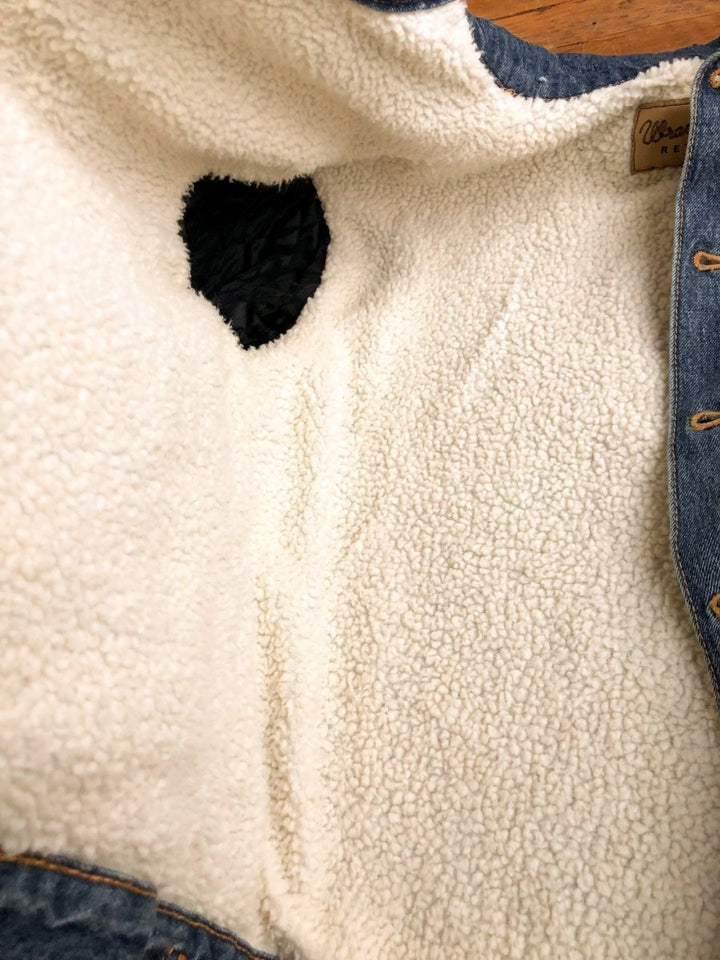 A close-up of The Wagoner men's jacket by Savannah Sevens western life{&}style, turned inside out to show white sherpa lining and a black patch near the armpit. The button-up is open and laid flat on a wooden surface.