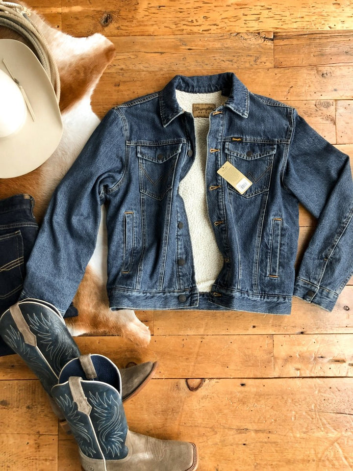 The Wagoner men's blue denim sherpa jacket by Savannah Sevens western life{&}style is displayed on a wooden floor beside a white cowboy hat, cowhide rug, and gray-blue cowboy boots with decorative stitching.
