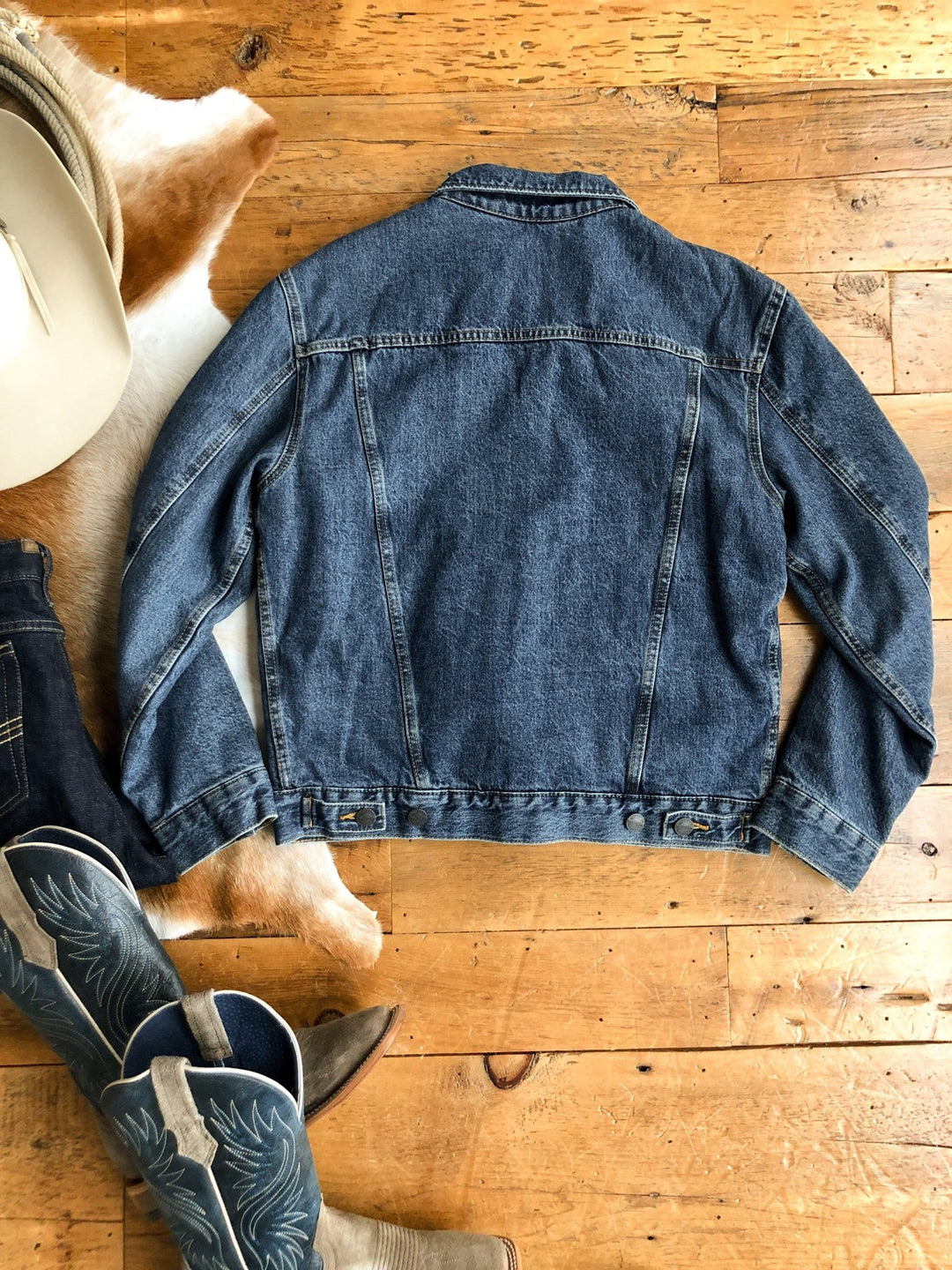 The Wagoner Men's button-up jacket by Savannah Sevens western life{&}style is laid flat, back facing up, on a wooden floor near cowboy boots, part of a cowboy hat, and a cowhide rug.