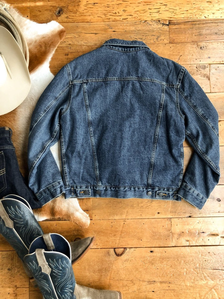 The Wagoner Men's button-up jacket by Savannah Sevens western life{&}style is laid flat, back facing up, on a wooden floor near cowboy boots, part of a cowboy hat, and a cowhide rug.
