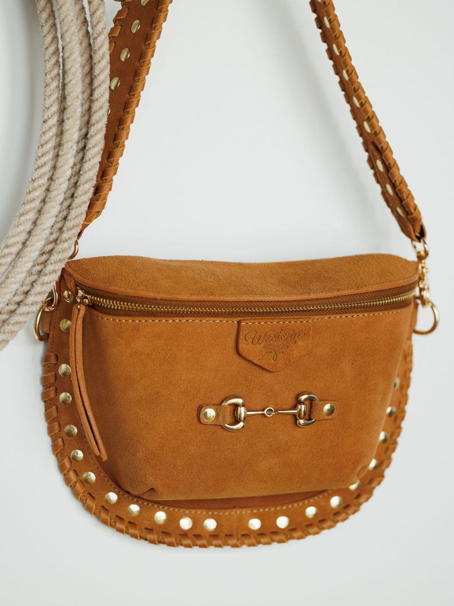 The Yoakum crossbody bag by Savannah Sevens western life{&}style, with tan suede, gold studs, snaffle bit detail, and stitched edges, hangs on a white wall. A looped rope in the background adds a Western vibe to the scene.