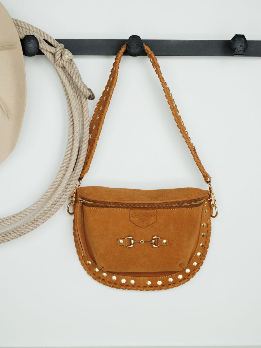 The Yoakum by Savannah Sevens western life{&}style—a tan suede shoulder bag with gold studs, braided strap, and gold hardware—hangs on a black wall hook beside a beige hat and coiled rope, channeling classic Western style.