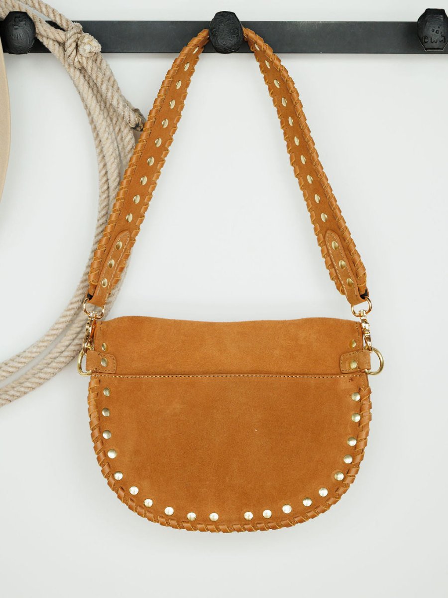 The Yoakum by Savannah Sevens western life{&}style—a tan suede handbag with a wide strap, gold studs, visible stitching, and snaffle bit accent—hangs on a black hook with a coiled rope against a white background. 