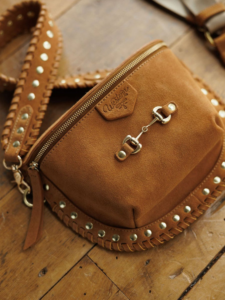 The Yoakum by Savannah Sevens western life{&}style is a brown suede crossbody with gold hardware, a snaffle bit detail on the strap adorned with gold studs, and stitched edging, displayed on a wooden surface.